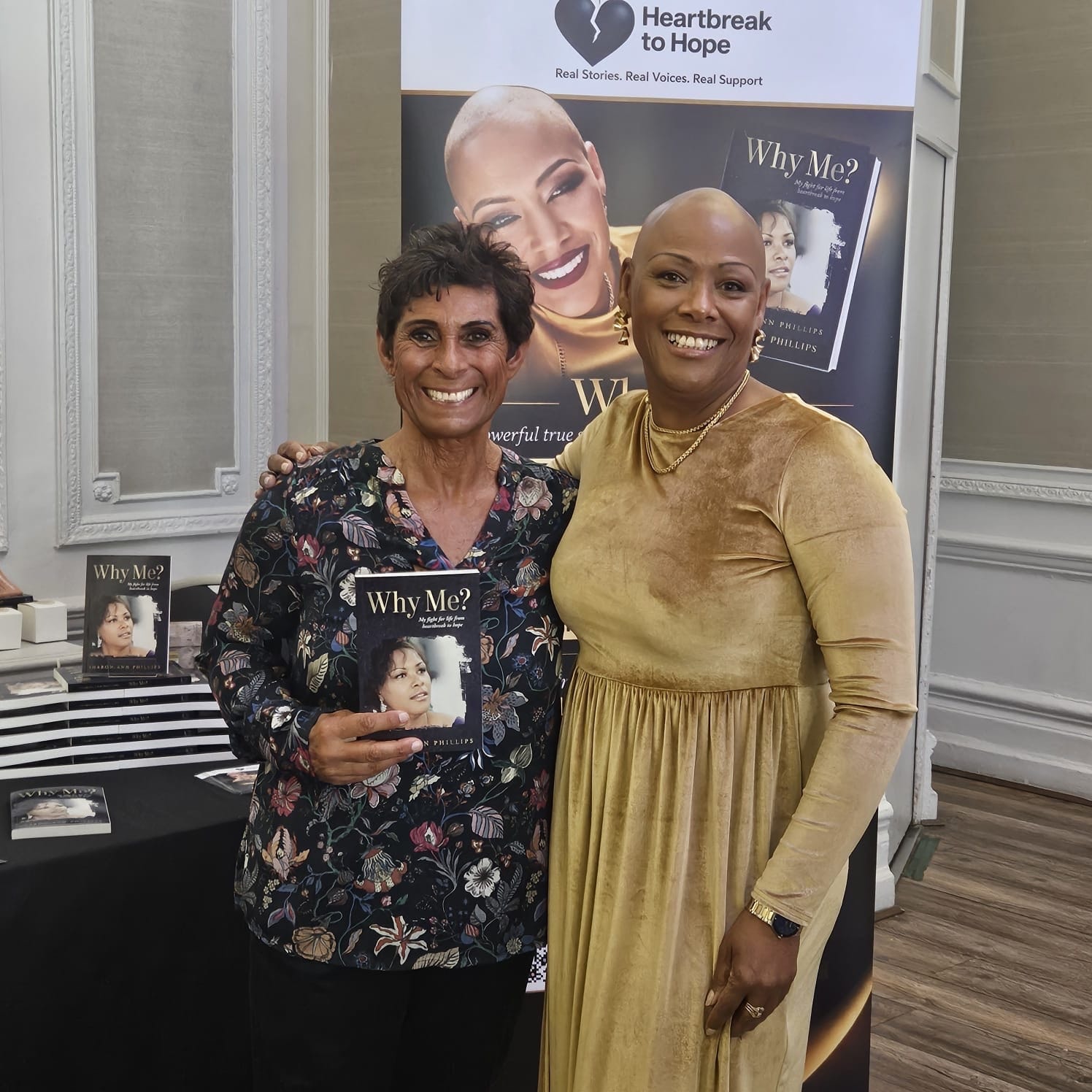 Sharon-Ann Phillips with Fatima Whitbread at a Heartbreak to Hope book event, both smiling in front of the Why Me? banner